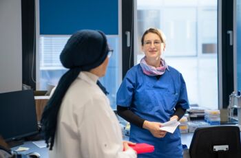 Zwei Frauen im Labor, eine in weißem Laborkittel und die andere in blauer Arbeitskleidung, unterhalten sich, während sie Laborproben und Unterlagen halten.