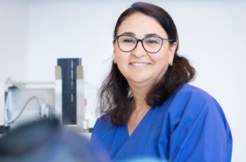 Auf dem Foto strahlt eine Frau mit dunklen Haaren und Brille in einem blauen Kittel voller Sympathie. Sie steht in einem Labor und lächelt vertrauensvoll in die Kamera.