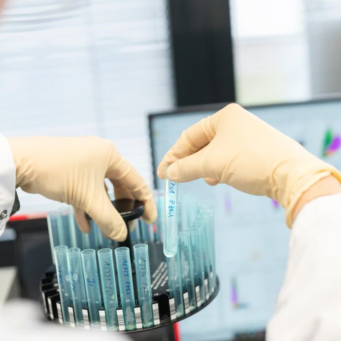 Probenanalyse in der Durchflusszytometrie Nahaufnahme zweier Hände in Einweghandschuhen, die Reagenzgläser in einem Halter arrangieren. Im Hintergrund ein Computerbildschirm mit Diagrammen zur Durchflusszytometrie.