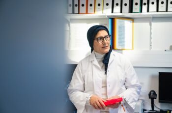 Frau in weißem Laborkittel mit Kopftuch hält einen roten Probenhalter, stehend vor Aktenregalen in einem Molekular-Laborraum.