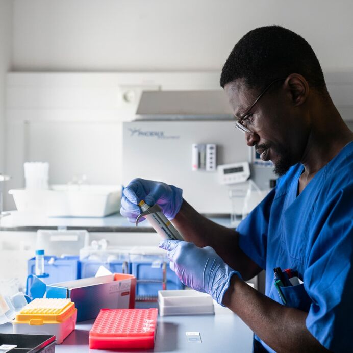 Volumen-Einstellung einer Pipette Mann in blauer Arbeitskleidung und Handschuhen arbeitet konzentriert mit einer Pipette an einem Labortisch, umgeben von Laborutensilien.