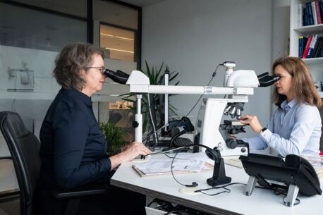 Zwei Frauen in einem modernen Labor sitzen sich gegenüber und arbeiten an einem Mikroskop. Eine Frau trägt ein dunkelblaues Oberteil, die andere ein hellblaues Hemd. Im Hintergrund sind Bücherregale und Pflanzen zu sehen.