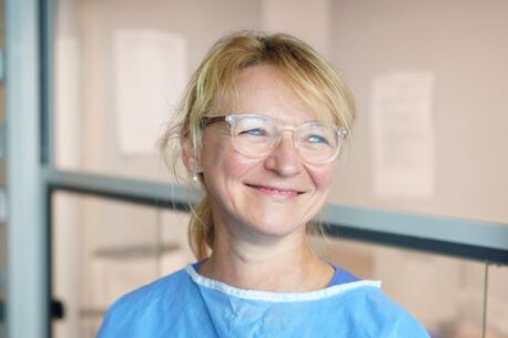 Portrait einer lächelnden Frau in blauer Schutzkleidung und Brille in einem Labor.