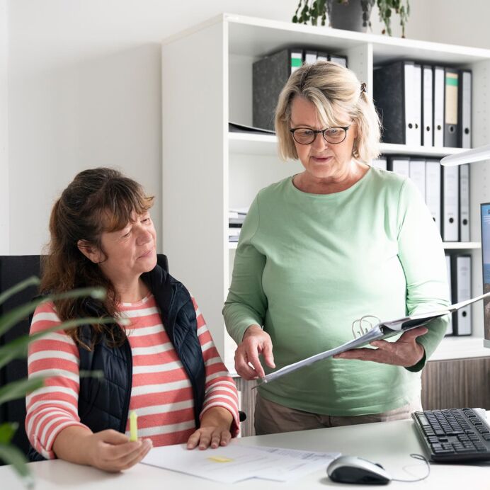 Unsere Ansprechpartnerinnen für Sie Zwei Kolleginnen arbeiten gemeinsam in einem Büro; eine sitzt und hört zu, während die andere steht und auf eine Mappe zeigt. Im Hintergrund befinden sich Regale mit Ordnern und ein Computerbildschirm.