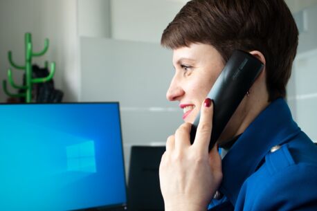 Diese Frau sitzt am Schreibtisch und hält einen Telefonhörer am Ohr, vor ihr ein Computerbildschirm. Sie trägt kurzes Haar.
