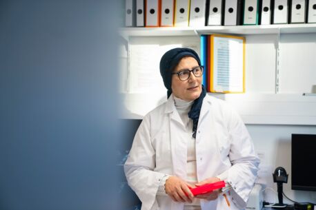 Frau in weißem Laborkittel mit Kopftuch hält einen roten Probenhalter, stehend vor Aktenregalen in einem Molekular-Laborraum.