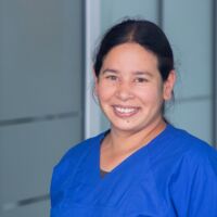 Dies ist ein Porträtfoto von Dr. Veronica Bernard. Sie trägt blaue Laborkleidung und blickt lächelnd in die Kamera. Ihre dunklen Haare sind zu einem Zopf gebunden.