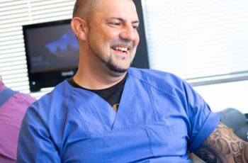 Auf dem Foto ist ein Mann im blauen Kittel abgebildet, der in einem Labor steht und fröhlich lacht. Er scheint die Arbeit gerade mit viel Freude und Engagement zu betreiben.