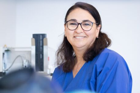 Auf dem Foto strahlt eine Frau mit dunklen Haaren und Brille in einem blauen Kittel voller Sympathie. Sie steht in einem Labor und lächelt vertrauensvoll in die Kamera.
