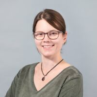 Eine Frau mit braunen Haaren und Brille trägt ein grünes Oberteil und eine Kette, steht lächelnd mit verschränkten Armen vor einem grauen Hintergrund.