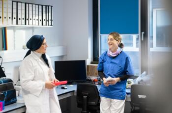 Zwei Frauen, eine im weißen Laborkittel und die andere in blauer Arbeitskleidung, stehen im Laborbüro und unterhalten sich, umgeben von Aktenordnern und Laborgeräten.