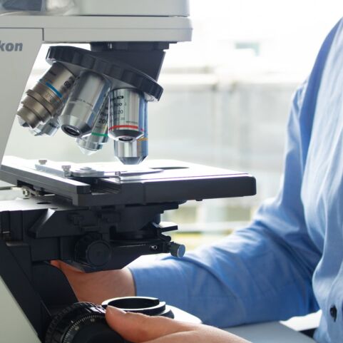 In dieser Nahaufnahme ist ein Mann im blauen Hemd am Mikroskop zu sehen, wobei nur ein Ausschnitt des Mannes sichtbar ist.