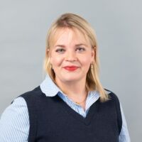 Eine blonde Frau trägt ein blau-weiß gestreiftes Hemd mit einem dunkelblauen Pullunder und steht lächelnd vor einem grauen Hintergrund.