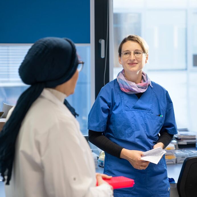 Labor-Besprechung Zwei Frauen im Labor, eine in weißem Laborkittel und die andere in blauer Arbeitskleidung, unterhalten sich, während sie Laborproben und Unterlagen halten.