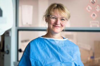 Portrait einer lächelnden Frau in blauer Schutzkleidung und Brille in einem Labor.