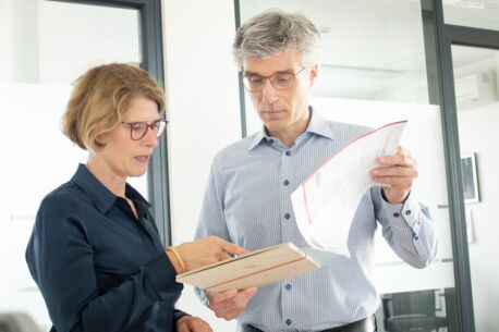 Die Frau und der Mann sind in eine angeregte Unterhaltung vertieft, wobei er eine Mappe in den Händen hält und sie mit dem Finger auf etwas in der Mappe zeigt.