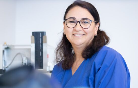 Auf dem Foto strahlt eine Frau mit dunklen Haaren und Brille in einem blauen Kittel voller Sympathie. Sie steht in einem Labor und lächelt vertrauensvoll in die Kamera.