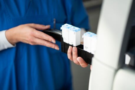 Hände einer Person in blauer Arbeitskleidung halten eine Halterung mit weißen Laborbehältern, die in ein Färbe-Automat eingesetzt werden.