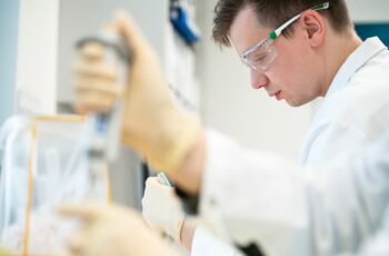 Mann in weißem Laborkittel und Schutzbrille arbeitet mit einer Pipette konzentriert im Labor.