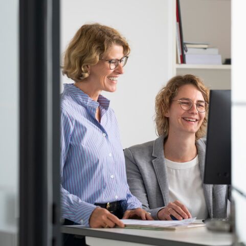 Ausbildung einer Weiterbildungsärztin Zwei Frauen in einem modernen Büro arbeiten gemeinsam am Computer. Die Frau links trägt ein gestreiftes Hemd, die rechte einen grauen Blazer. Beide wirken konzentriert und lächeln, während sie an einem Bildschirm arbeiten.