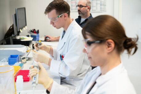 Zwei Labormitarbeiter in weißen Kitteln und Schutzbrillen arbeiten mit Pipetten an Proben, während ein Kollege im Hintergrund an einem Computer sitzt.
