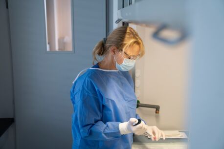 Frau in blauer Schutzkleidung, Maske und Handschuhen arbeitet konzentriert mit einer Pipette im Labor.