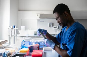 Mann in blauer Arbeitskleidung und Handschuhen arbeitet konzentriert mit einer Pipette an einem Labortisch, umgeben von Laborutensilien.