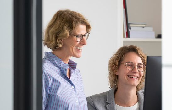Zwei Frauen in einem modernen Büro arbeiten gemeinsam am Computer. Die Frau links trägt ein gestreiftes Hemd, die rechte einen grauen Blazer. Beide wirken konzentriert und lächeln, während sie an einem Bildschirm arbeiten.
