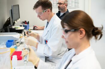 Zwei Labormitarbeiter in weißen Kitteln und Schutzbrillen arbeiten mit Pipetten an Proben, während ein Kollege im Hintergrund an einem Computer sitzt.