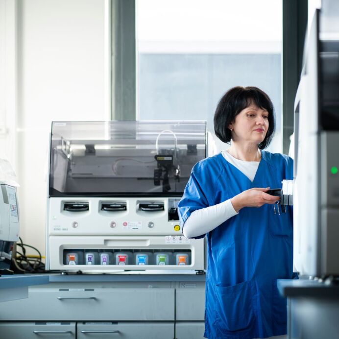 Bestücken der Färbeautomaten Frau in blauer Arbeitskleidung arbeitet an einem modernen Analysegerät in einem Laborraum mit weiteren Laborgeräten im Hintergrund.