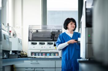 Frau in blauer Arbeitskleidung arbeitet an einem modernen Analysegerät in einem Laborraum mit weiteren Laborgeräten im Hintergrund.