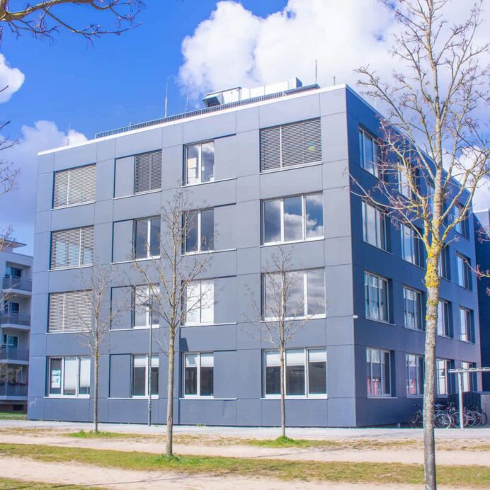 Hämatopathologie Lübeck - Das Institutsgebäude Dieses Foto zeigt die Außenansicht eines Gebäudekomplexes eines Instituts, umgeben von vielen Bäumen. Der Himmel ist blau und leicht bewölkt..