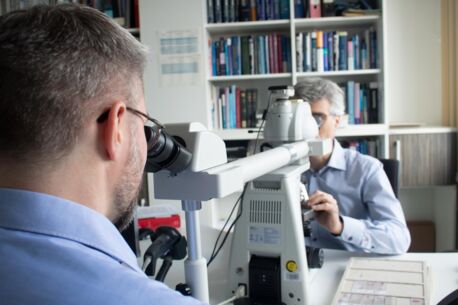 Auf dem Foto sehen wir zwei Männer am Mikroskop sitzen, die sich gegenüber sitzen. Im Hintergrund ein großes Bücherregal.