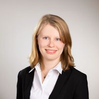 Porträtfoto einer jungen Frau mit blonden Haaren, weißer Bluse und schwarzem Blazer vor neutralem Hintergrund.