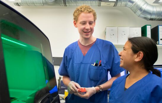 Auf dem Foto sehen wir einen Mann und eine Frau in blauen Kitteln in einem Labor stehen. Sie betrachten sich gegenseitig und scheinen sich über etwas zu freuen. Links sieht man ein großes, technisches Gerät.