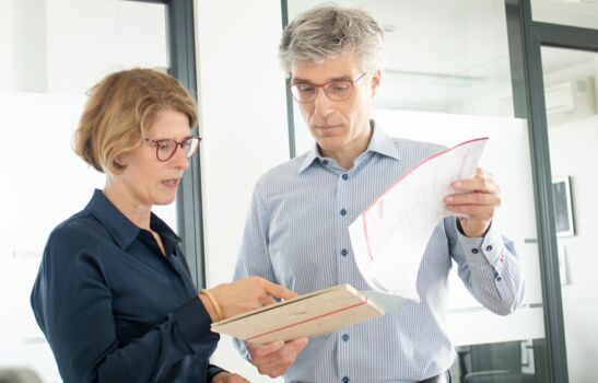 Die Frau und der Mann sind in eine angeregte Unterhaltung vertieft, wobei er eine Mappe in den Händen hält und sie mit dem Finger auf etwas in der Mappe zeigt.