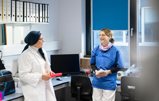Zwei Mitarbeiterinnen der Hämatopathologie Lübeck unterhalten sich im Labor: links eine Frau im weißen Laborkittel mit dunklem Kopftuch und rotem Probenhalter, rechts eine Frau in blauer Arbeitskleidung mit Unterlagen in der Hand.