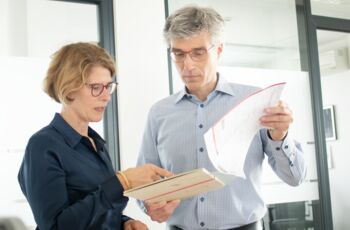 Die Frau und der Mann sind in eine angeregte Unterhaltung vertieft, wobei er eine Mappe in den Händen hält und sie mit dem Finger auf etwas in der Mappe zeigt.